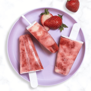 purple kids plate on a marble kitchen counter with three strawberry popsicles with swirled greek yogurt on the plate with sliced strawberries.