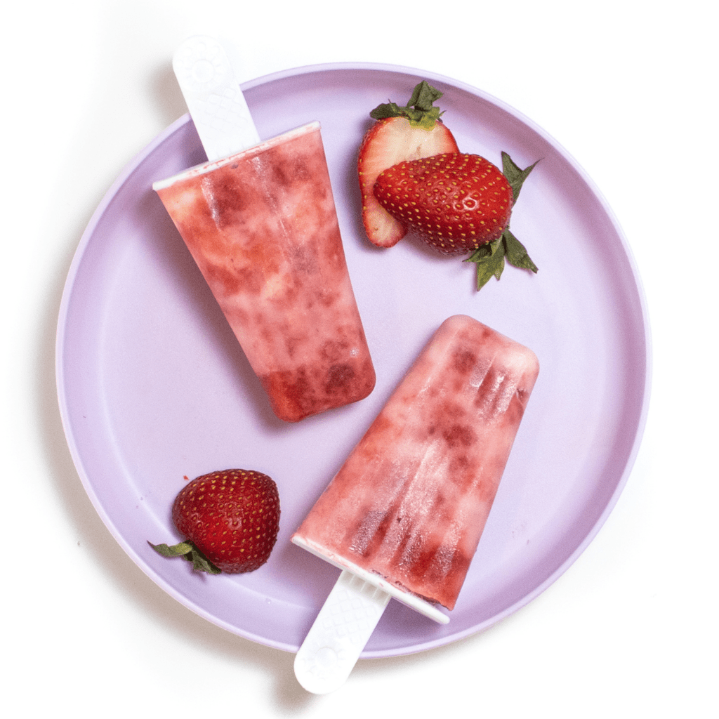 A purple color plate with two swirled strawberry yogurt popsicles and sliced strawberries against a white background.