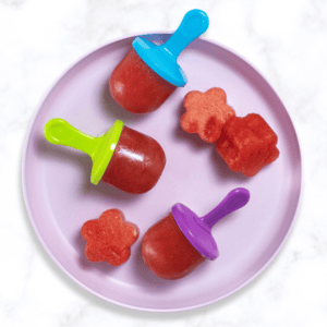 purple kids plate with 3 small watermelon popsicles and a few fun cut out shapes of watermelon. The plate is sitting on my white marble kitchen table.