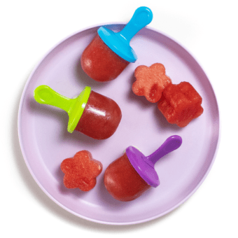 Purple toddler plate with three watermelon popsicles with colorful popsicle sticks and chunks of watermelon on the plate against a white background.