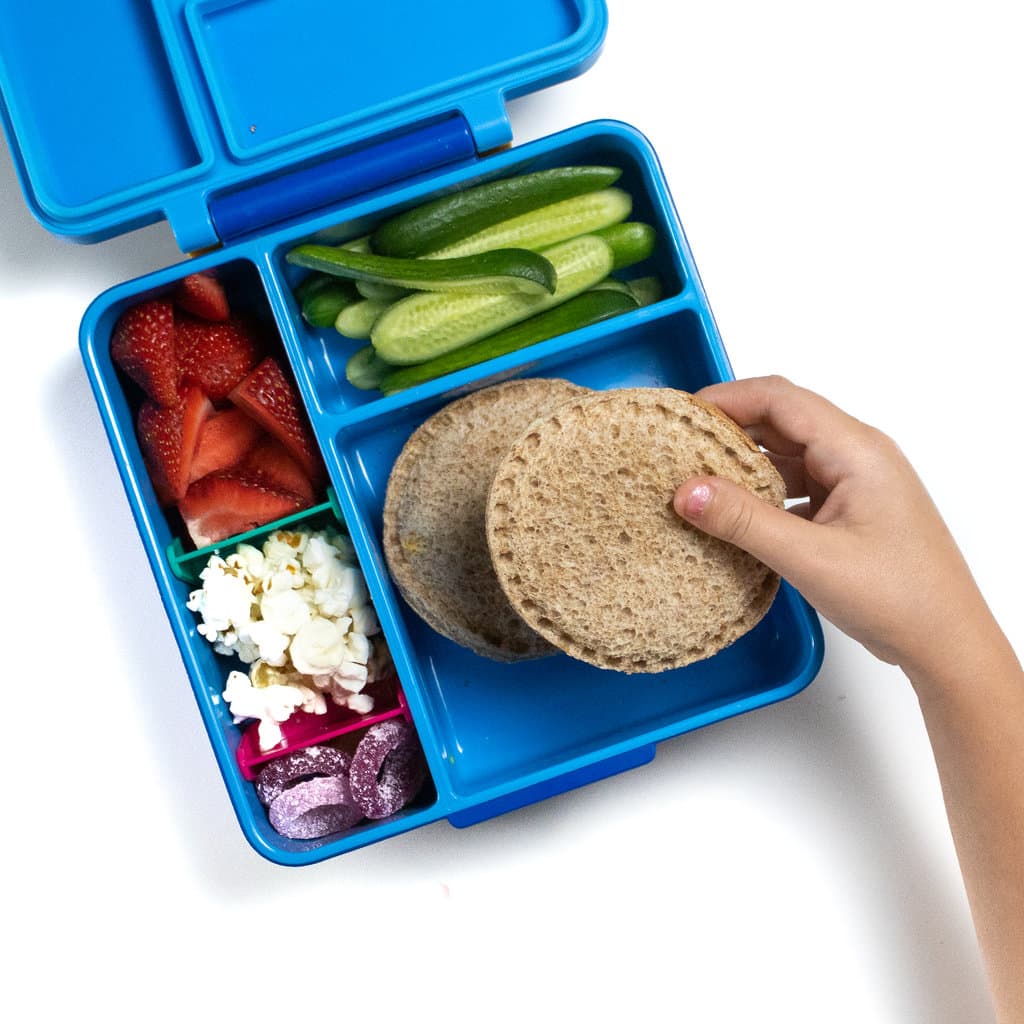 A blue lunchbox filled with two uncrustables sandwiches, cucumbers, strawberries, popcorn and gummy snacks, with a small kids hand holding one of the sandwiches.