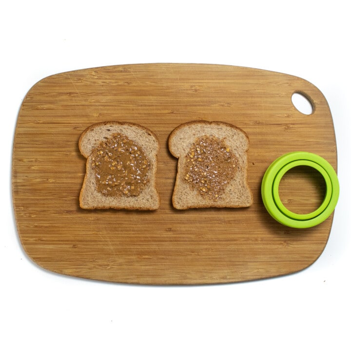 A cutting board with two pieces of bread and an uncrustable cutter.