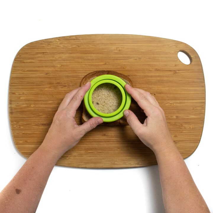 Two hands holding a sandwich cutter over two pieces of bread on a wooden cutting board.