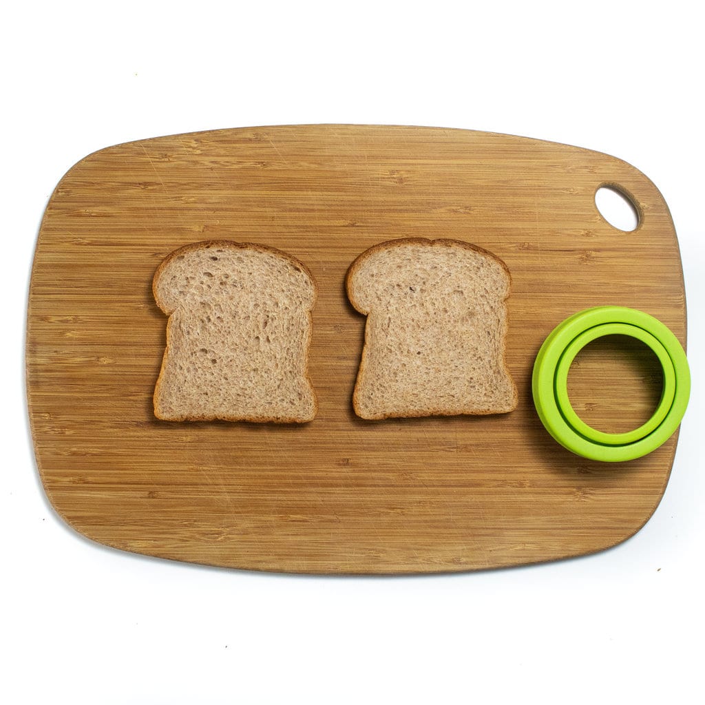 Two pieces of bread on a wooden cutting board with a green uncrustable sandwich cutter. 