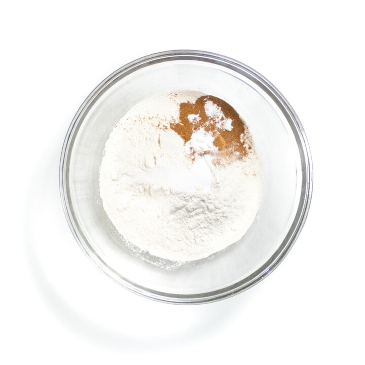 A clear mixing bowl against a white background with flour, cinnamon and baking powder.