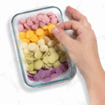A small glass container with five different flavors of baby yogurt melts. The container is sitting on a marble countertop with my hand reaching over and grabbing an mango carrot flavor melt.