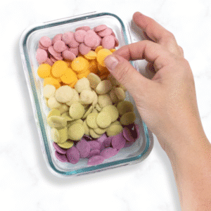 A small glass container with five different flavors of baby yogurt melts. The container is sitting on a marble countertop with my hand reaching over and grabbing an mango carrot flavor melt.