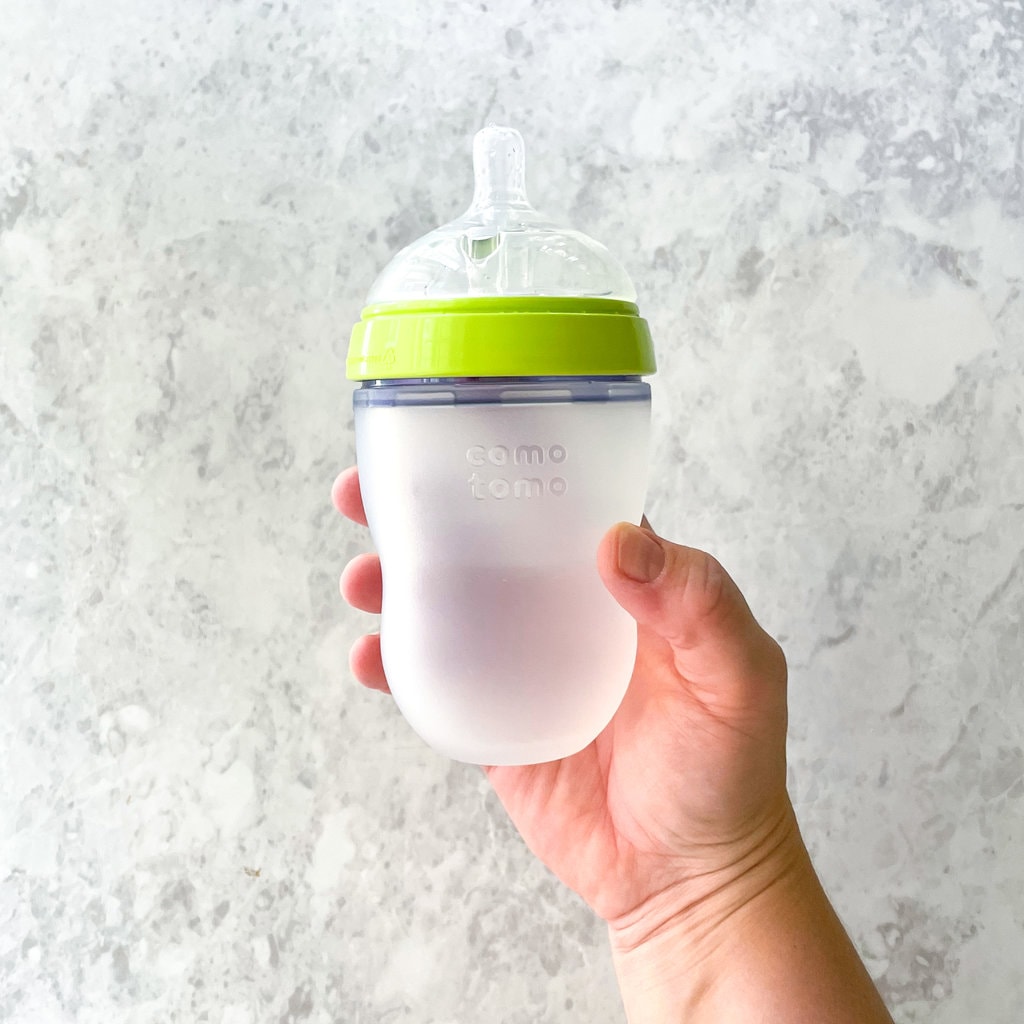 A hand holding a comotomo baby bottle against a white marble countertop.