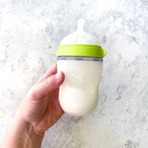 A hand holding a comotomo baby bottle against a white marble countertop.