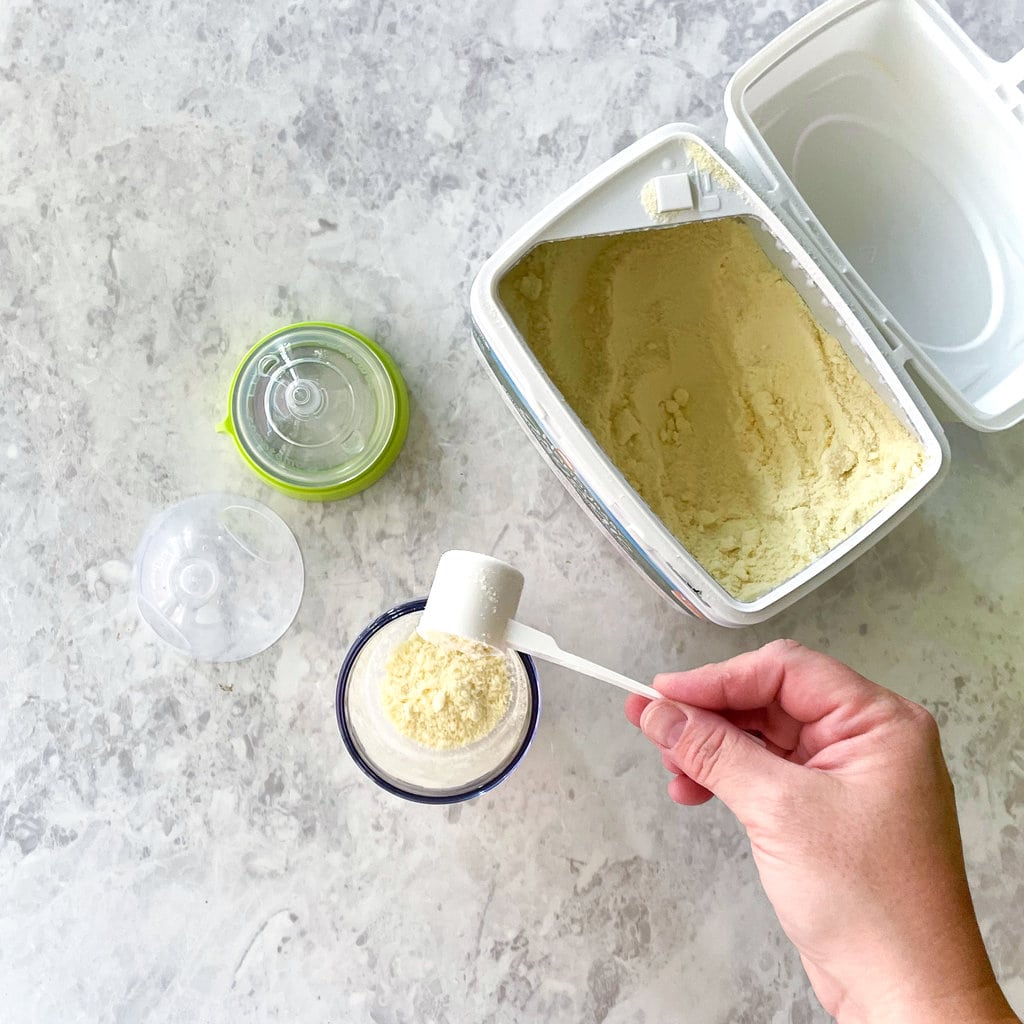 Hand scooping formula into a Comotomo baby bottle against a marble countertop.