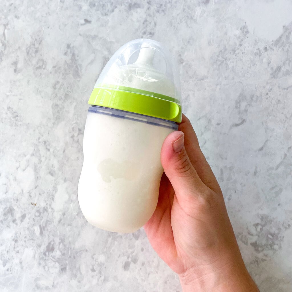 a comotomo bottle full of formula with a hand holding it against a marble countertop.