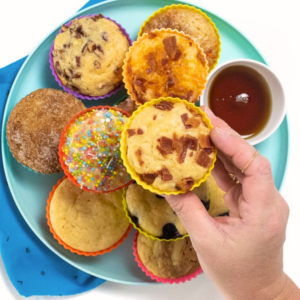 My hand holding a pancake muffin over a teal plate full of other pancake muffins with various topping.