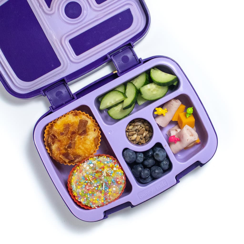A purple lunchbox with pancake muffins with two different toppings, cut cucumbers, blueberries and turkey roll ups.