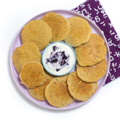 A purple kids plate full of zucchini pancakes with a small bowl of yogurt and blueberry sauce I can see a white background with a purple napkin.