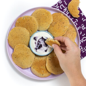 A purple kids plate full of zucchini pancakes with a small bowl of yogurt and blueberry sauce I can see a white background with a purple napkin with a hand holding a pancake dipping it into the yogurt sauce.