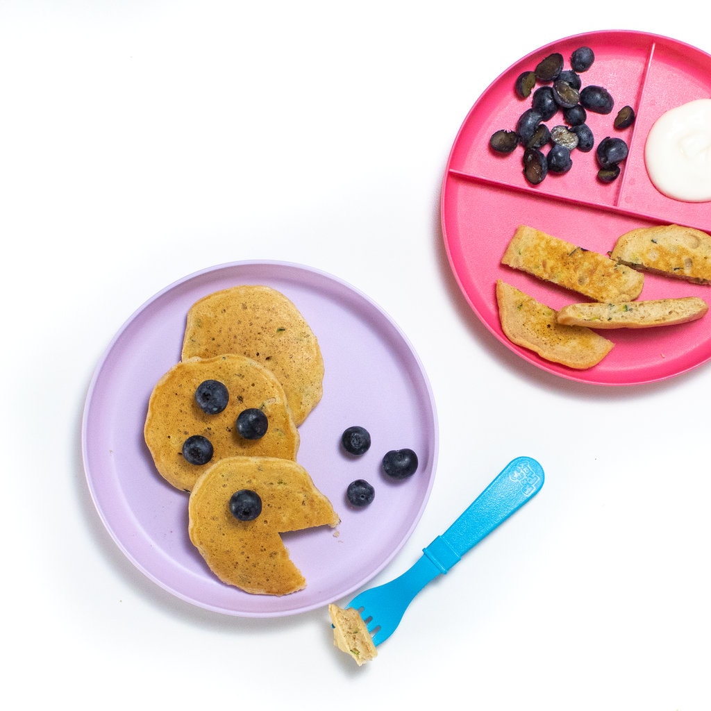 Two kids played against a white background with zucchini pancakes cut into several different ways with blueberries, and yogurt.
