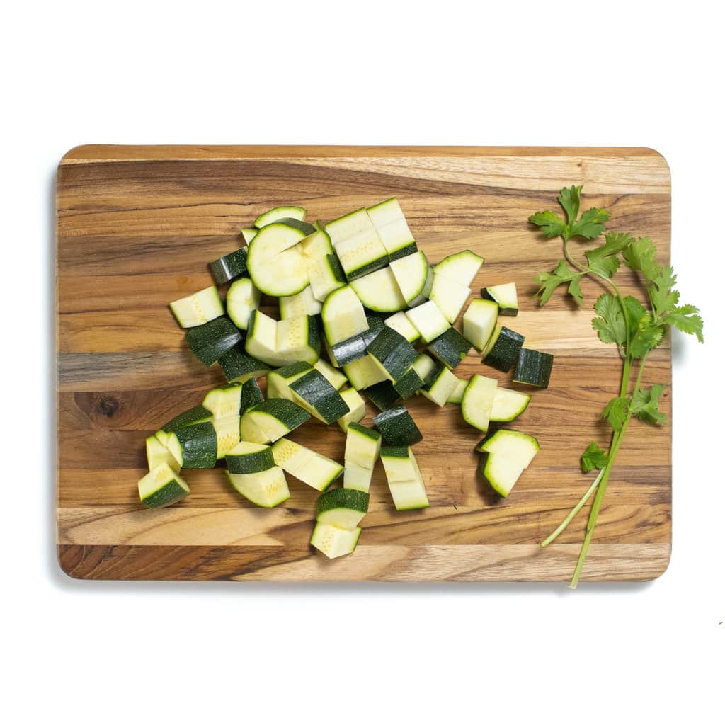 A cutting board with cut zucchini and a few sprigs of cilantro against a white background.