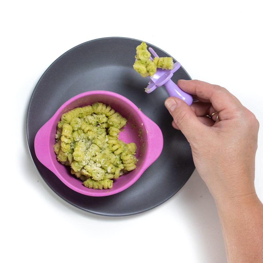 A gray plate with a pink baby bowl on top filled with a chunky zucchini broccoli pasta for Baby, with a hand holding a purple baby spoon with pasta on it.