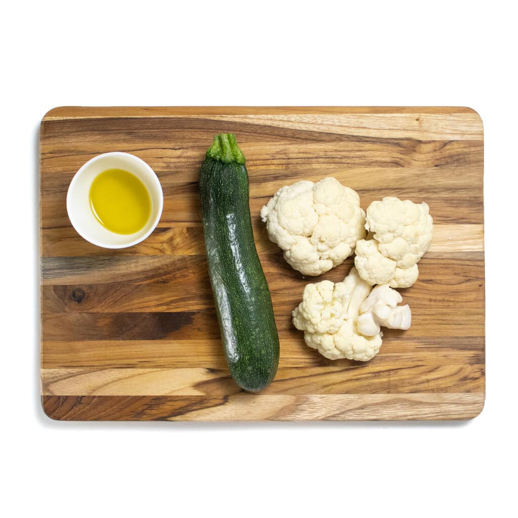 A wooden cutting board against a white background with a zucchini, cauliflower and olive oil.