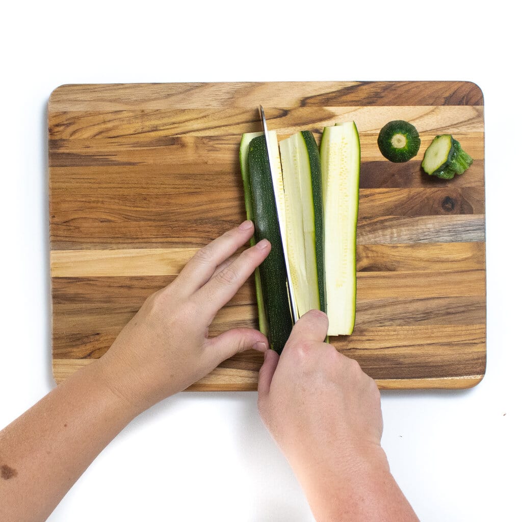 Two hands cutting a zucchini into small pieces on a wooden cutting board against a white background.
