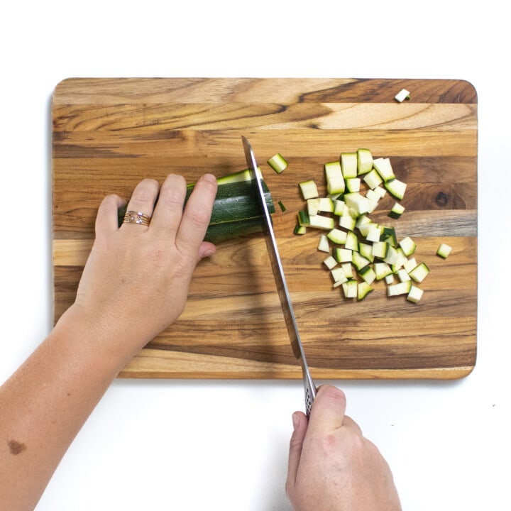Two hands cutting a zucchini into small pieces on a wooden cutting board against a white background.