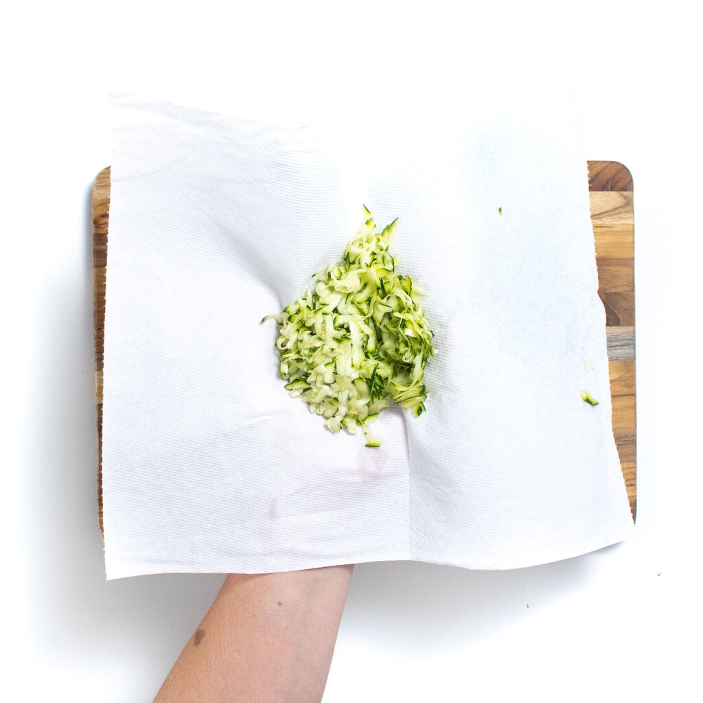 A hand holding a paper towel with grated zucchini in it.