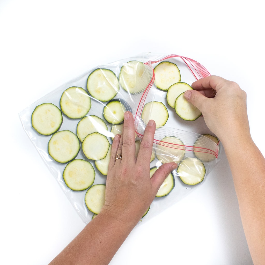 2 Hands placing in slices of fresh zucchini into a Ziploc freezer bag.