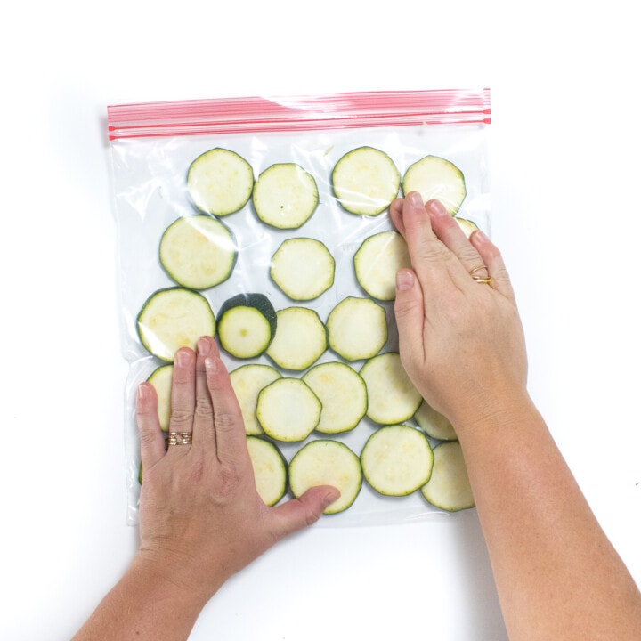 Two hands pressing the air out of a Ziploc bag full of sliced zucchini.