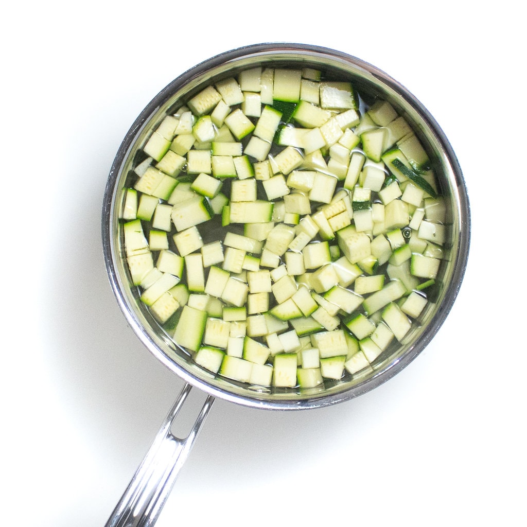 A silver sauce pan full of diced zucchini with water that is boiling.