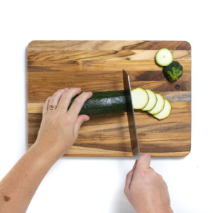 A wooden cutting board with two hands cutting a zucchini into slices.
