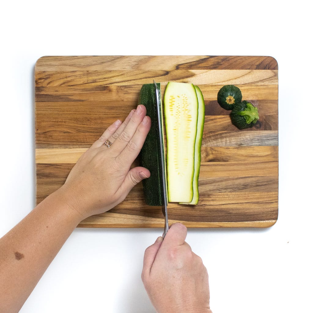A wooden cutting board with two hands cutting a zucchini into planks.
