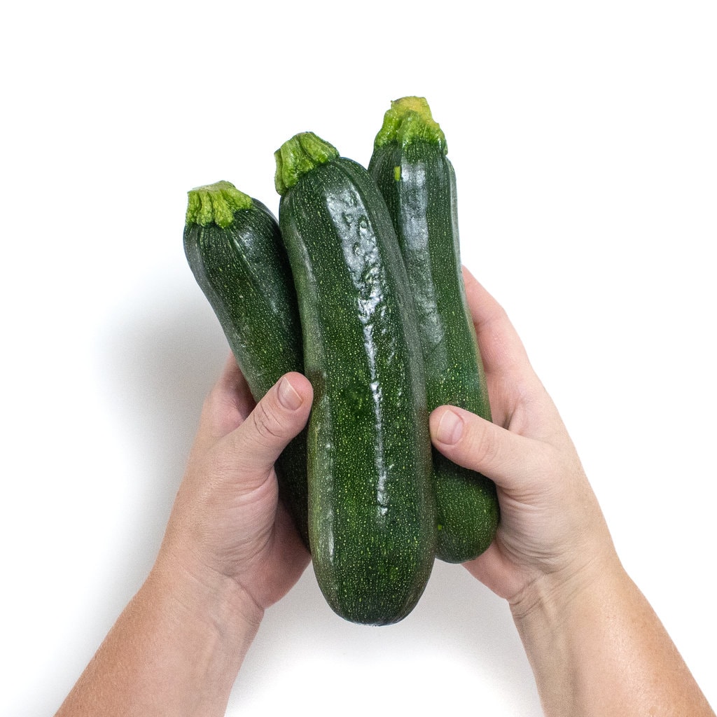 Hands holding three zucchini against a white background.