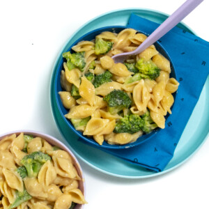 A blue kids plate and bowl with a blue napkin and purple fork, with broccoli mac & cheese inside the bowl. Next to this is a pink bowl full of broccoli mac & cheese.
