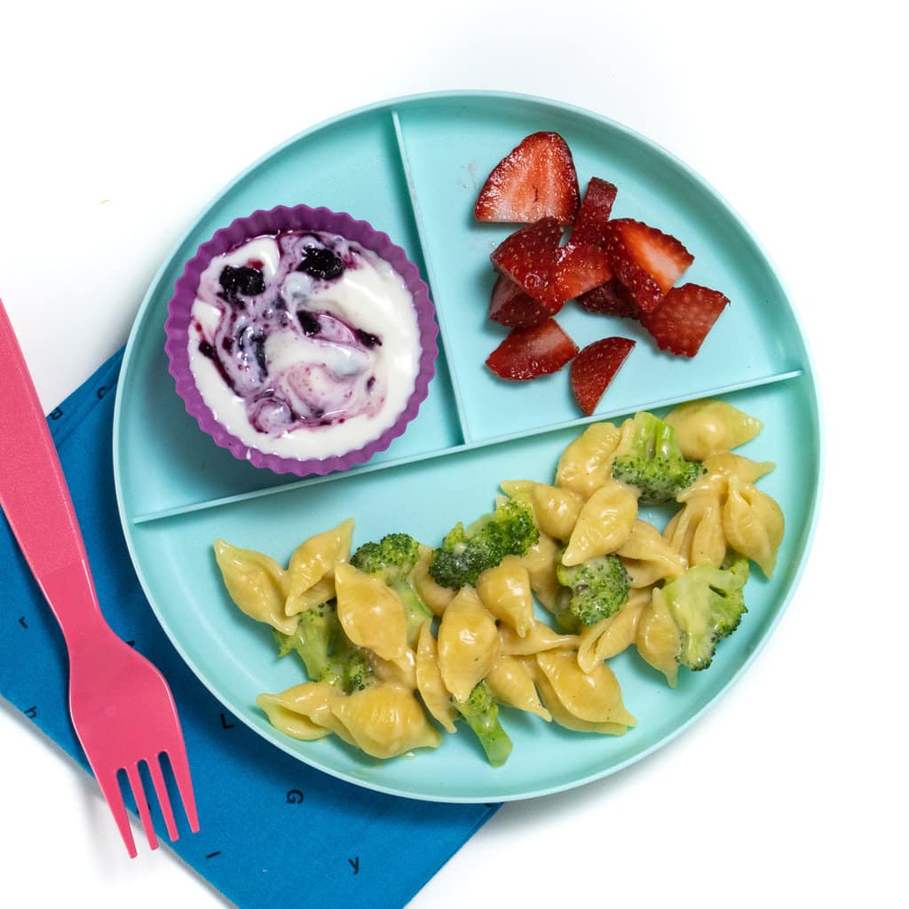 Kids three section plate with broccoli mac & cheese, sliced strawberries and yogurt against a white background with a blue napkin and pink fork.