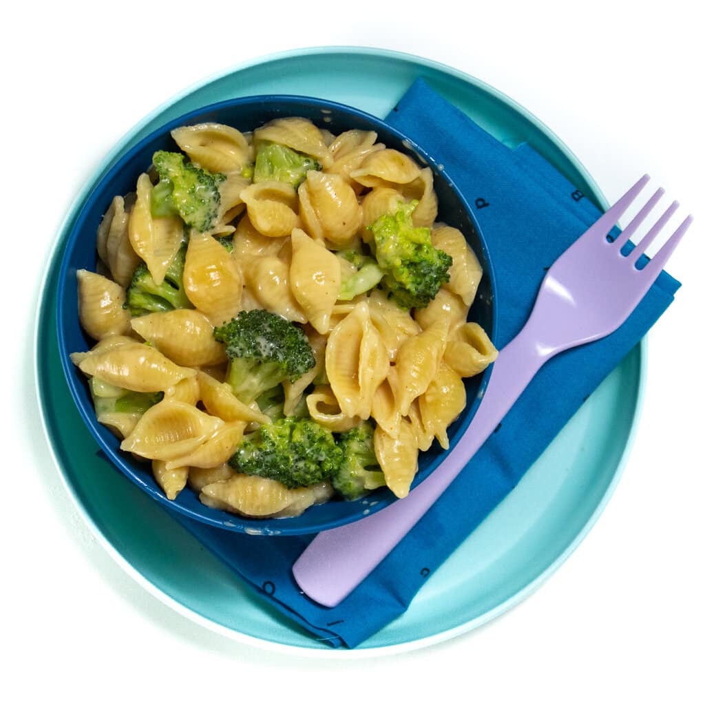 A blue plate and a blue bowl full of broccoli mac & cheese with a purple fork with mac & cheese on it against a white background with a blue napkin.