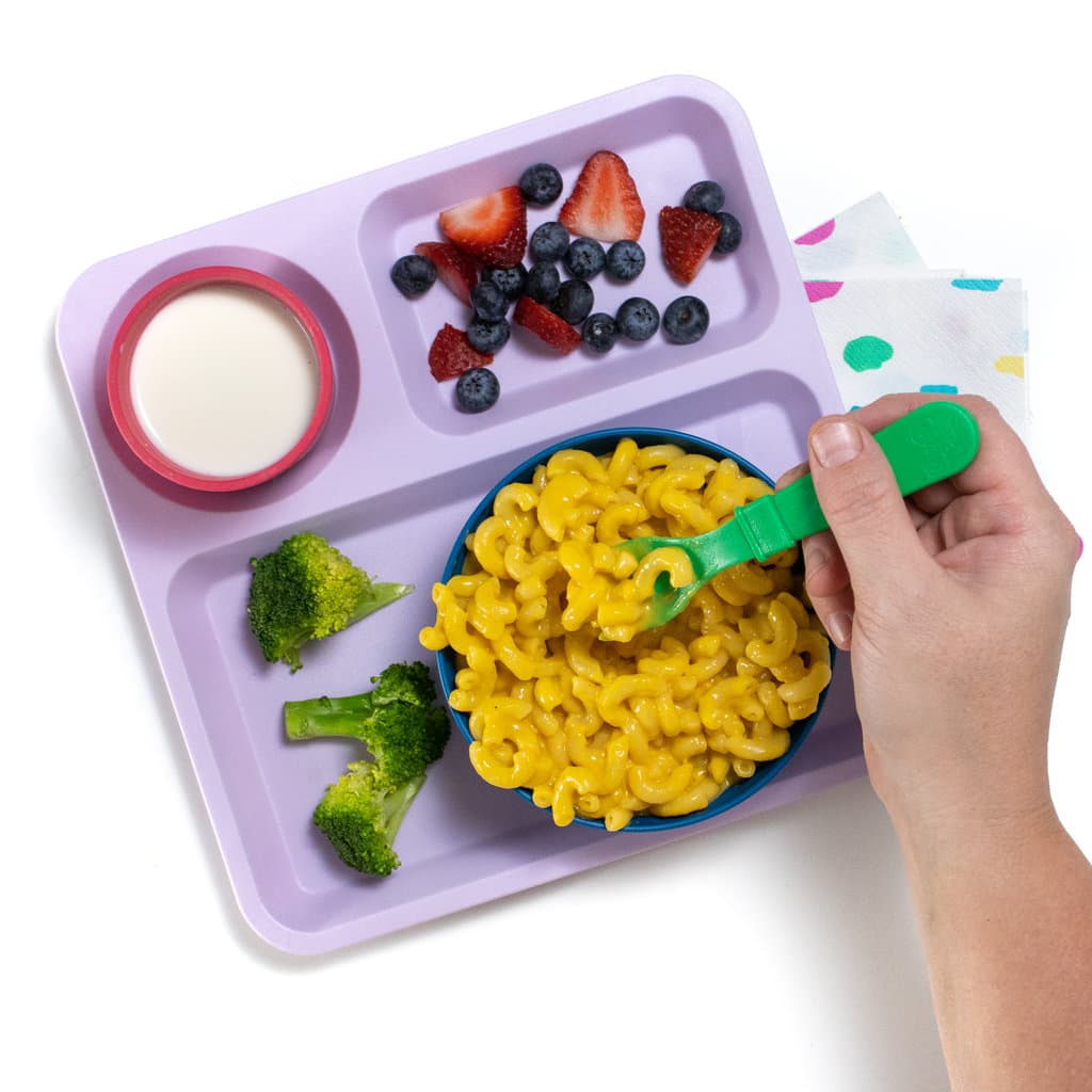 A purple kids plate with a bowl full of butternut squash mac & cheese, broccoli, berries and milk. Along with a colorful napkin and green fork against a white background.