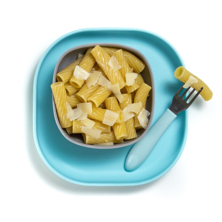 Gray baby bowl sitting on a blue baby plate with tube pasta and flakes of cheese on top.
