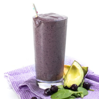 A side view of a pregnancy smoothie that is purple with a purple napkin and ingredients scattered at the bottom.