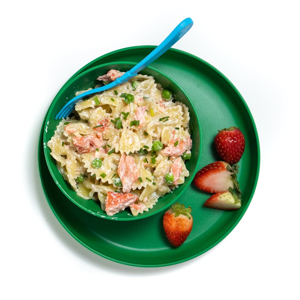 Green plate and bowl full of salmon pasta and peas with strawberries on the side.