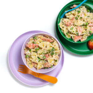 Set of purple and green kids bowls and plates with salmon pasta with peas inside.
