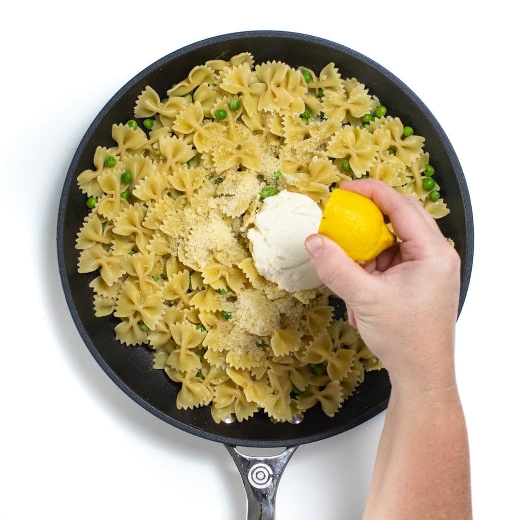 Hand squeezing a lemon over a skilled filled with pasta, peas, ricotta and parmesan cheese.