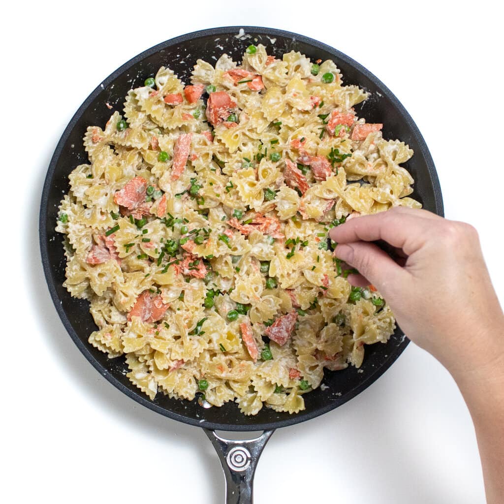 Skillet with pasta, peas, salmon and a hand reaching in and sprinkling chives on top.