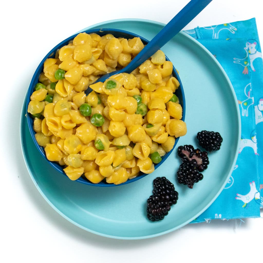 A dark blue kids plate on top of a light blue kids plate with a fun blue napkin, the bowl is full of sweet potato macaroni and cheese with peas.