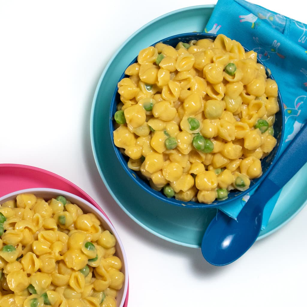 Fun colored kids plates and bowls full of shell pasta made into sweet potato macaroni and cheese with peas.