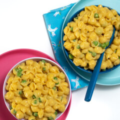 Fun colored kids plates and bowls full of shell pasta made into sweet potato macaroni and cheese with peas.