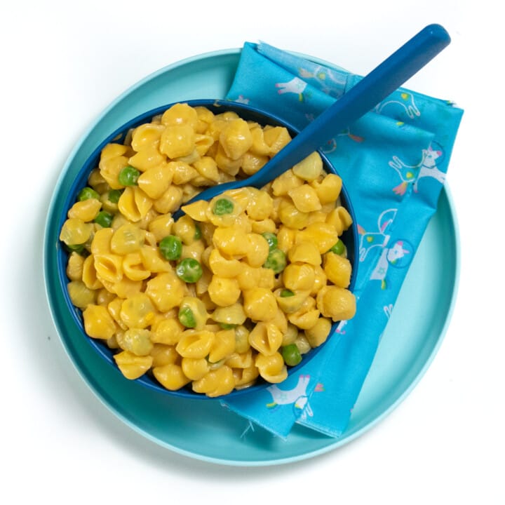 A dark blue kids plate on top of a light blue kids plate with a fun blue napkin, the bowl is full of sweet potato macaroni and cheese with peas.