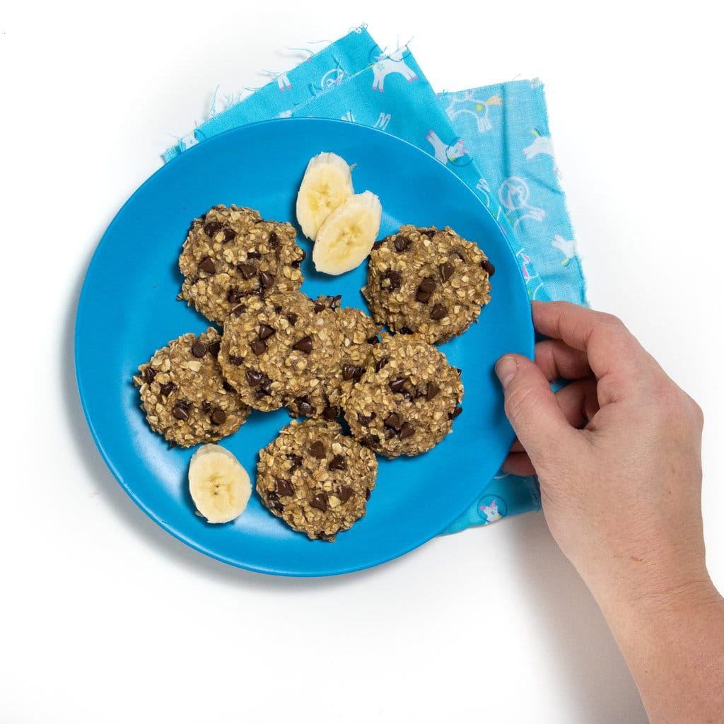 Hand holding a banana oatmeal chocolate chip cookie above a plate of cookies against a white background with the blue tablecloth.