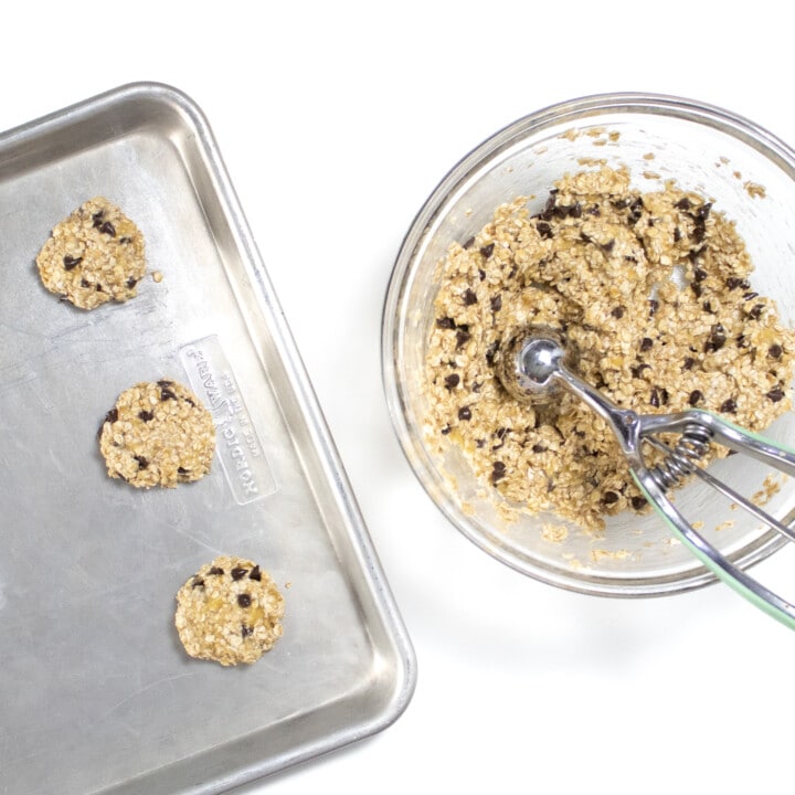 Clear glass bowl with banana cookies being scooped onto a baking sheet against the way back drop