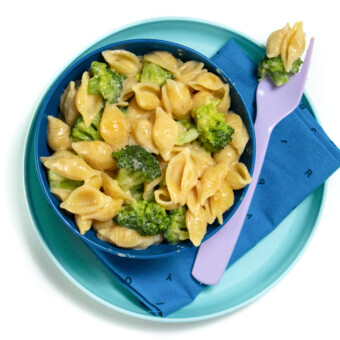 A blue plate and a blue bowl full of broccoli mac & cheese with a purple fork with mac & cheese on it against a white background with a blue napkin.