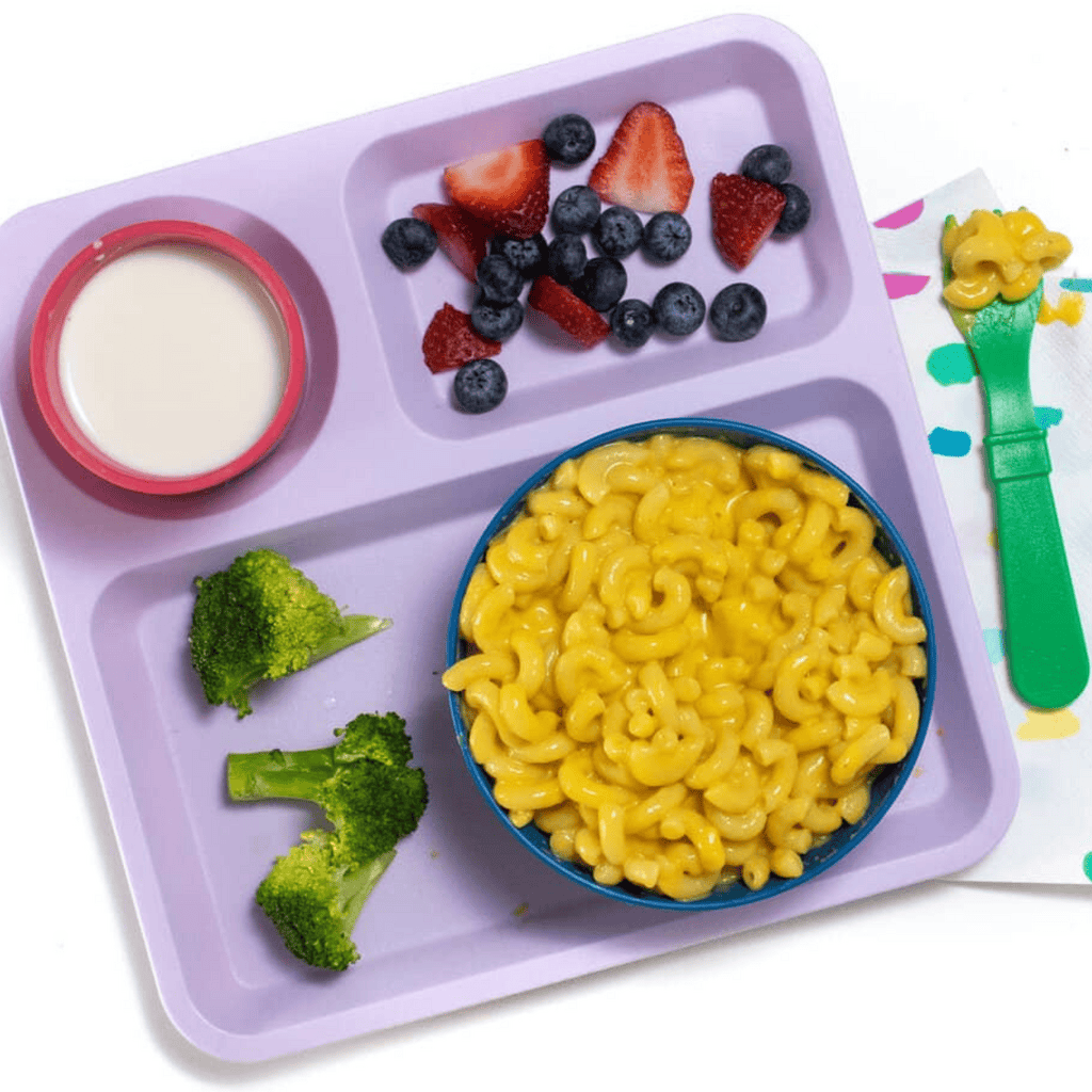 My hand holding a green kids spoon over a bowl of creamy butternut squash mac and cheese. The mac and cheese is in a blue kids bowl sitting on a school purple plate with a side of broccoli and fruit, all sitting on my white marble kitchen counter.